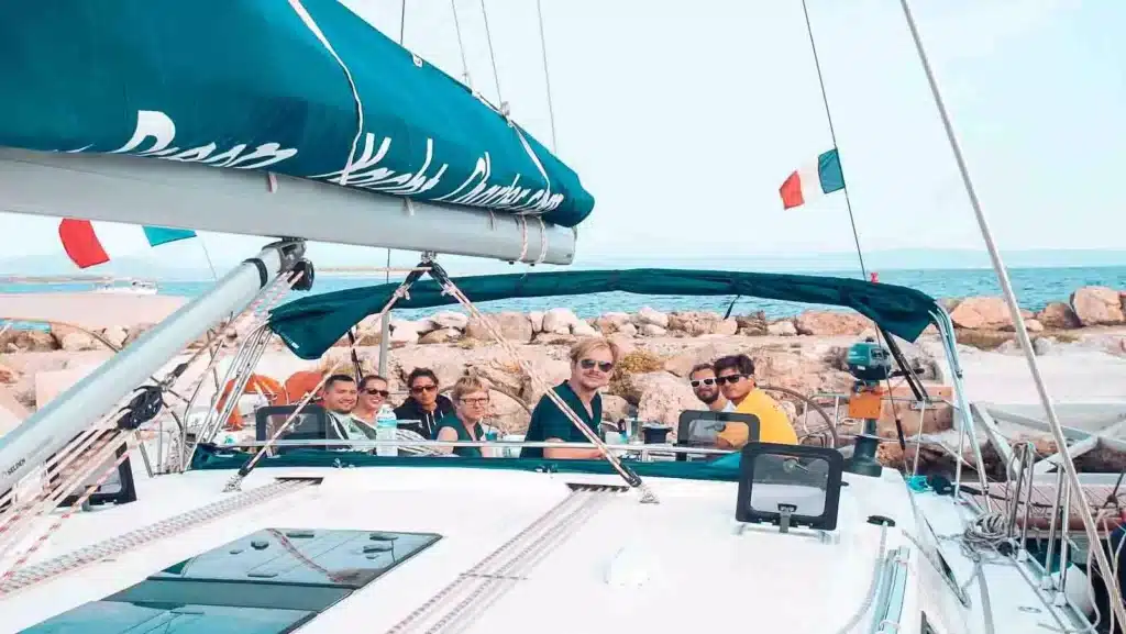 Eine Gruppe von sechs lächelnden Menschen auf einer Segelyacht, nahe einer felsigen Küste unter klarem Himmel. Auf dem Verdeck des Bootes steht „Dolce Vita“.