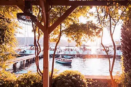 Ein ruhiger Blick auf den See bei Sonnenuntergang, eingerahmt von einer mit Weinreben umrankten Holzpergola, mit Blick auf angedockte Boote, darunter auch Segelyachten, die sanft auf dem glitzernden Wasser schaukeln.