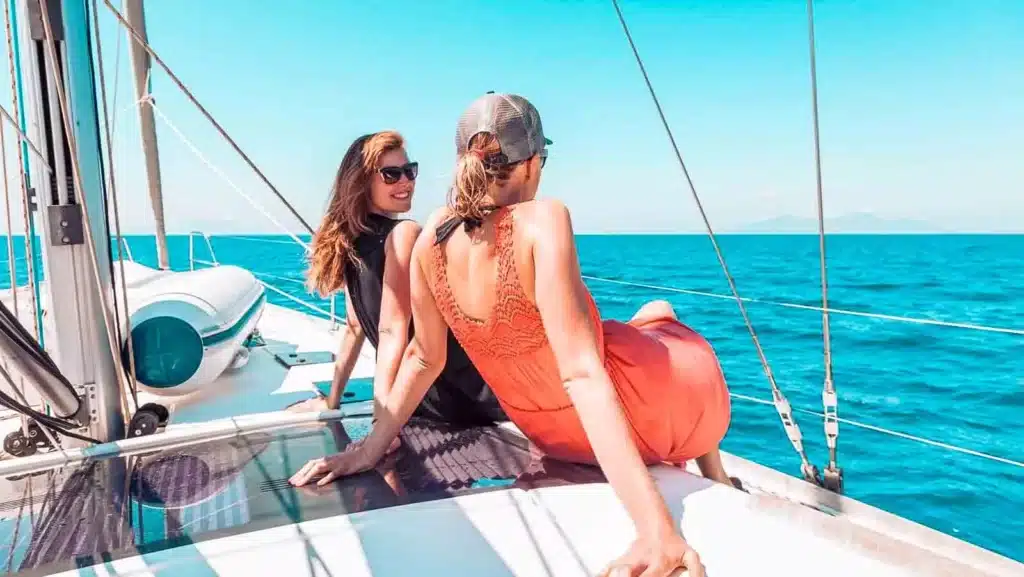 Zwei Frauen in Sommerkleidung genießen die Aussicht, während sie während eines Segeltörns auf dem Deck eines Segelboots sitzen, mit klarem blauen Himmel und dem Meer im Hintergrund.