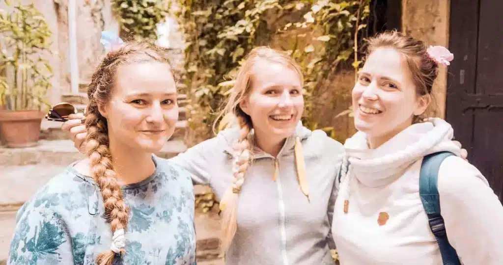 Drei Frauen mit fröhlichen Gesichtern posieren zusammen in einer gemütlichen Gasse während eines Segeltörns. Zwei Frauen haben geflochtene Haare, die mit Schleifen geschmückt sind, und eine andere trägt legere Kleidung und hat eine Kamera dabei.