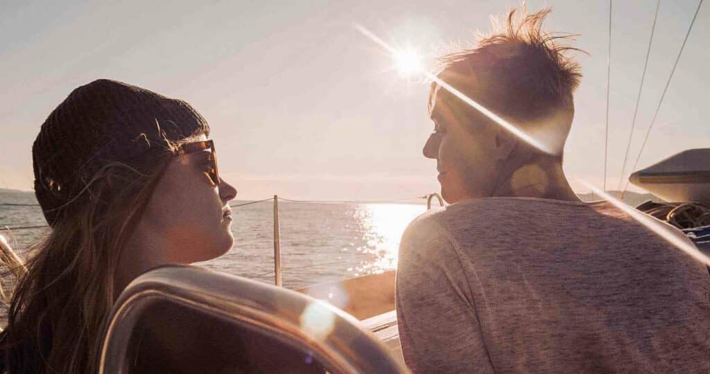 Zwei Menschen auf einer Segelreise bei Sonnenuntergang, die einander intim gegenüberstehen, während die Sonne zwischen ihnen hindurchscheint und ihre Silhouetten hervorhebt.