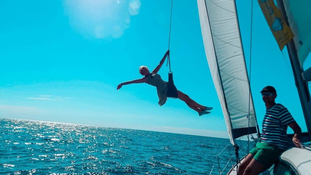 Eine Person schwingt an einem Seil auf einem Segelboot, das über dem Meer unter einem klaren blauen Himmel ausgestreckt ist. Eine andere Person, die ein gestreiftes Hemd und eine Sonnenbrille trägt, beobachtet vom Deck aus während ihres Segelt