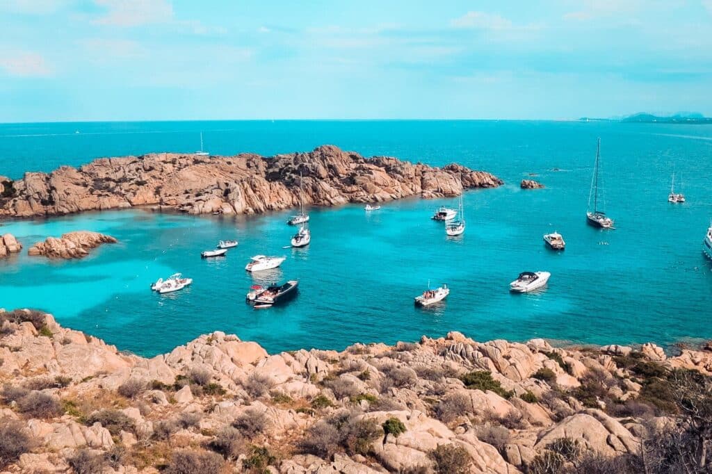 Eine malerische Küstenansicht mit türkisfarbenem Wasser, umgeben von rötlichen Felsformationen, mit mehreren Booten, die in Ufernähe unter einem klaren blauen Himmel vor Anker liegen, perfekt für einen Segelurlaub.