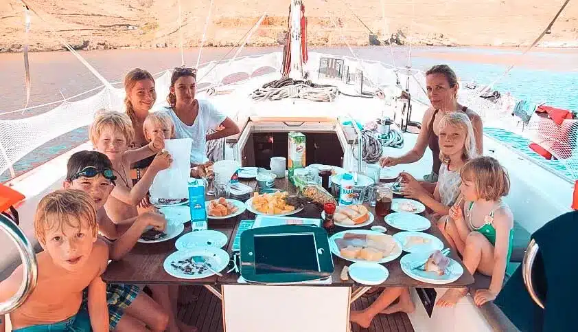 Eine Gruppe von zehn Personen, darunter sechs Kinder und vier Erwachsene, genießt eine Mahlzeit auf einer Segelyacht mit malerischer Flusskulisse. Sie lächeln und sitzen um einen mit Essen gefüllten Tisch herum.