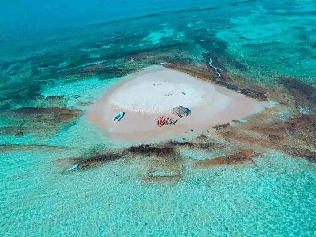 Luftaufnahme einer kleinen Sandinsel, umgeben von flachem, türkisfarbenem Wasser. Ein paar Menschen genießen auf ihrer Segelreise diesen abgeschiedenen Fleck mitten im Ozean.