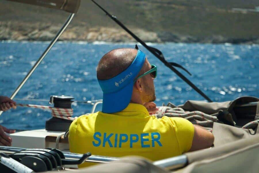 Ein Mann mit einem blauen Stirnband und einem gelben T-Shirt mit der Aufschrift „Skipper“ steuert eine Segelyacht mit einem klaren blauen Meer und einer zerklüfteten Küste im Hintergrund.