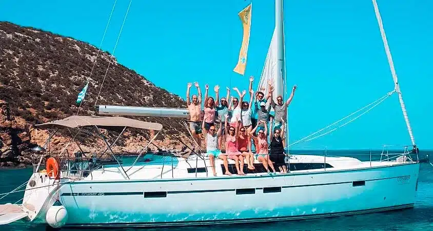Eine Gruppe fröhlicher Menschen heben an einem sonnigen Tag auf einer Segelyacht mit klarem, blauem Wasser und Hügeln im Hintergrund ihre Hände zum Jubeln.