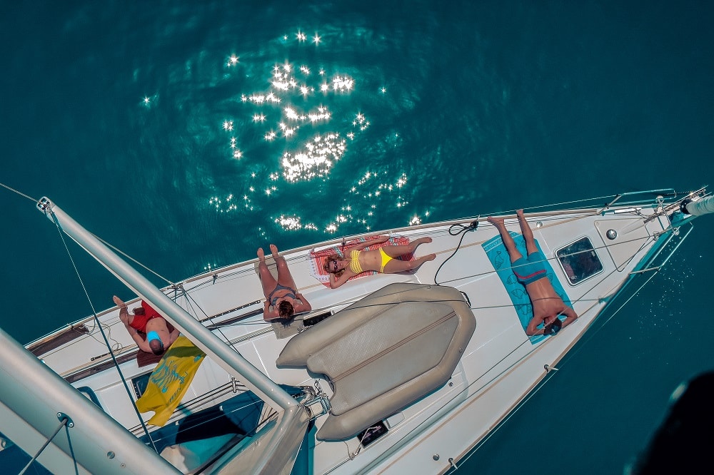 Luftaufnahme von vier Personen, die während eines Segelurlaubs auf dem Deck eines Segelboots ein Sonnenbad nehmen, das auf dem klaren, türkisfarbenen Wasser schwimmt, auf dessen Oberfläche sich ein Lichtmuster spiegelt.
