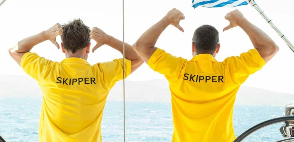 Zwei Männer in gelben „Skipper“-Hemden stehen am Steuer einer Segelyacht und blicken auf das Meer, wobei einer von ihnen das Steuerrad des Schiffs festhält.