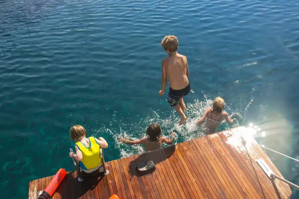 Ein Junge springt vom Deck eines Segelboots ins Wasser. Drei andere Kinder beobachten ihn, zwei weitere sind im Begriff, ihm nachzuspringen. Dies ist ein Paradebeispiel für eine freudige Sommeraktivität.