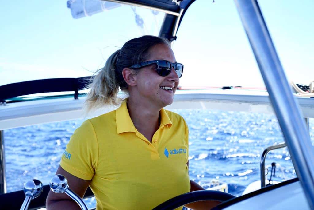 Eine Frau mit Sonnenbrille und gelbem Hemd lächelt, während sie eine Segelyacht steuert, mit klarem blauen Himmel und Meer im Hintergrund.