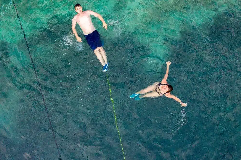 Luftaufnahme von zwei Personen, die im klaren, türkisfarbenen Wasser treiben, wobei eine Person an einem gelben Seil einer Segelyacht hängt.