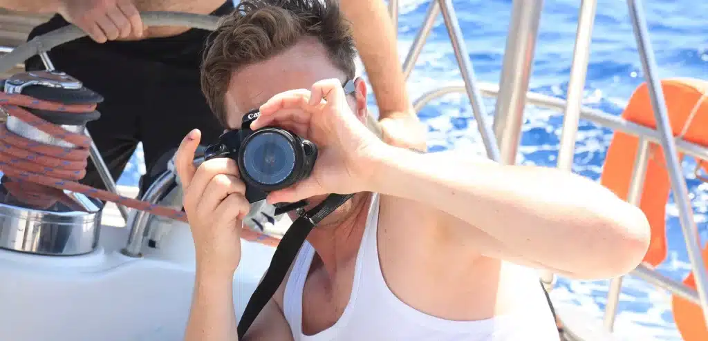Ein Mann mit einer Kamera macht an Bord einer Segelyacht ein Foto und fängt sein eigenes Bild durch die Linse ein, umgeben von nautischer Ausrüstung und dem blauen Ozean.