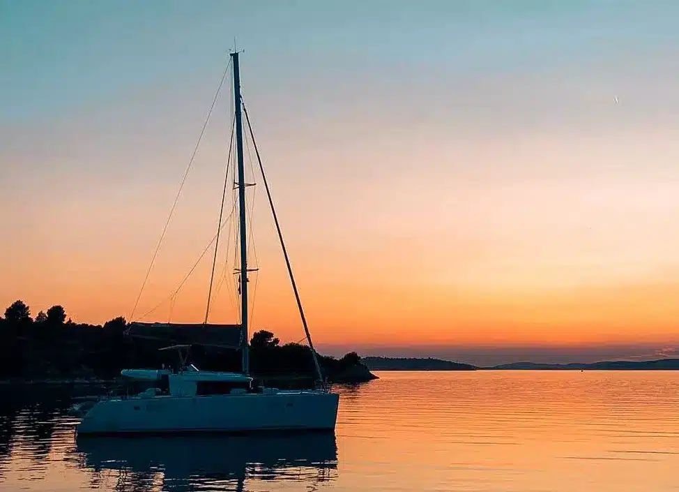 Eine Segelyacht, die auf ruhigen Gewässern vor einem Sonnenuntergangshimmel mit orangefarbenen und rosa Farbtönen verankert ist, die sich sanft im Wasser spiegeln.