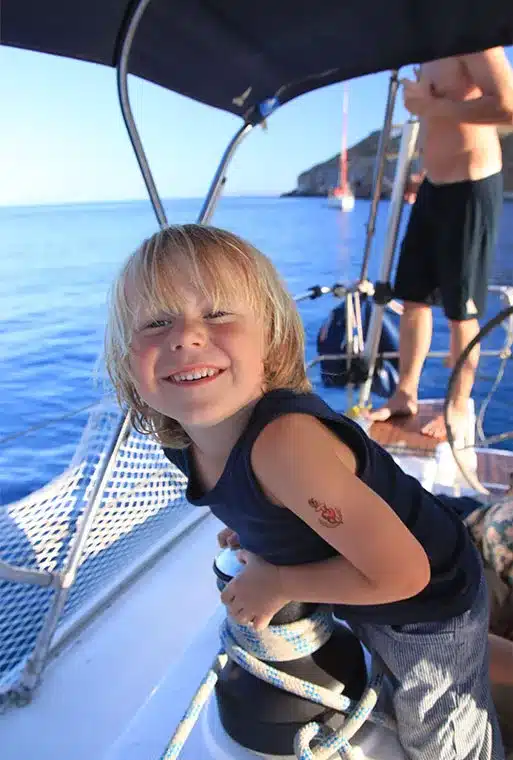 Ein kleiner Junge mit blondem Haar lächelt fröhlich auf einer Segelyacht, mit einem klaren blauen Himmel und Meer im Hintergrund und einer anderen Person, die teilweise hinter ihm sichtbar ist.