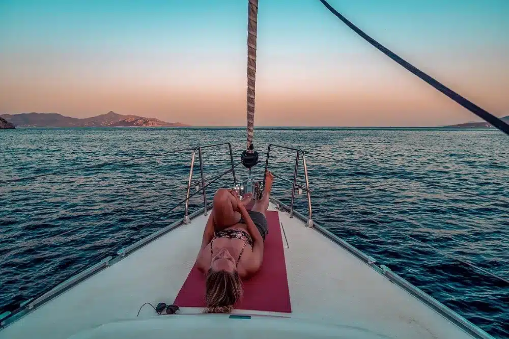 Eine Person entspannt sich während einer Segelreise auf dem Bug eines Segelboots und genießt den heiteren Sonnenuntergang über dem ruhigen Ozean, in der Ferne zeichnen sich die Berge vor einem pastellfarbenen Himmel ab.