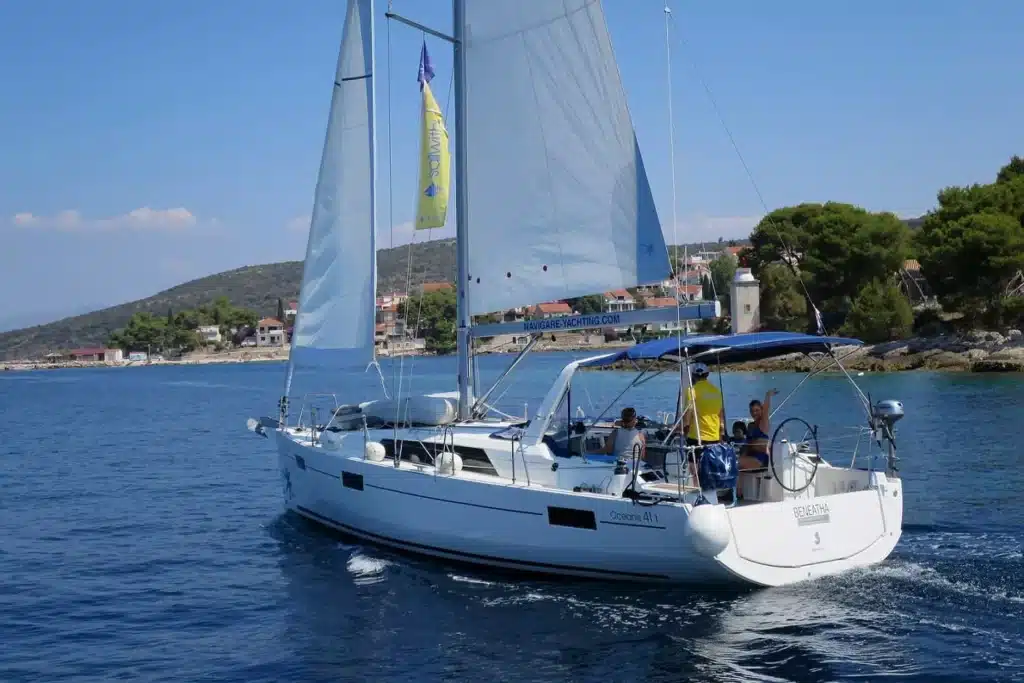 Ein weißes Segelboot mit voll ausgefahrenen Segeln kreuzt auf einem ruhigen blauen Meer in der Nähe einer Küstenregion mit Grünflächen und Gebäuden im Hintergrund. Auf dem Boot sind mehrere Personen zu sehen, die genießen