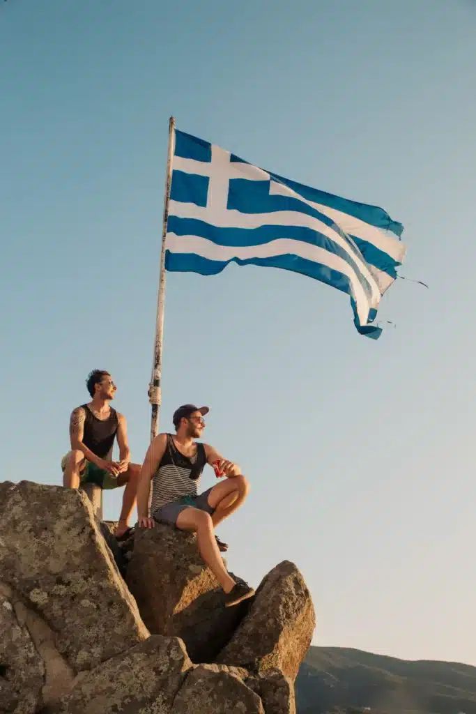 Zwei Menschen sitzen auf einem Felsen neben einer großen griechischen Flagge an einer Stange und blicken von ihrer Segelyacht auf eine malerische Aussicht mit klarem Himmel im Hintergrund.