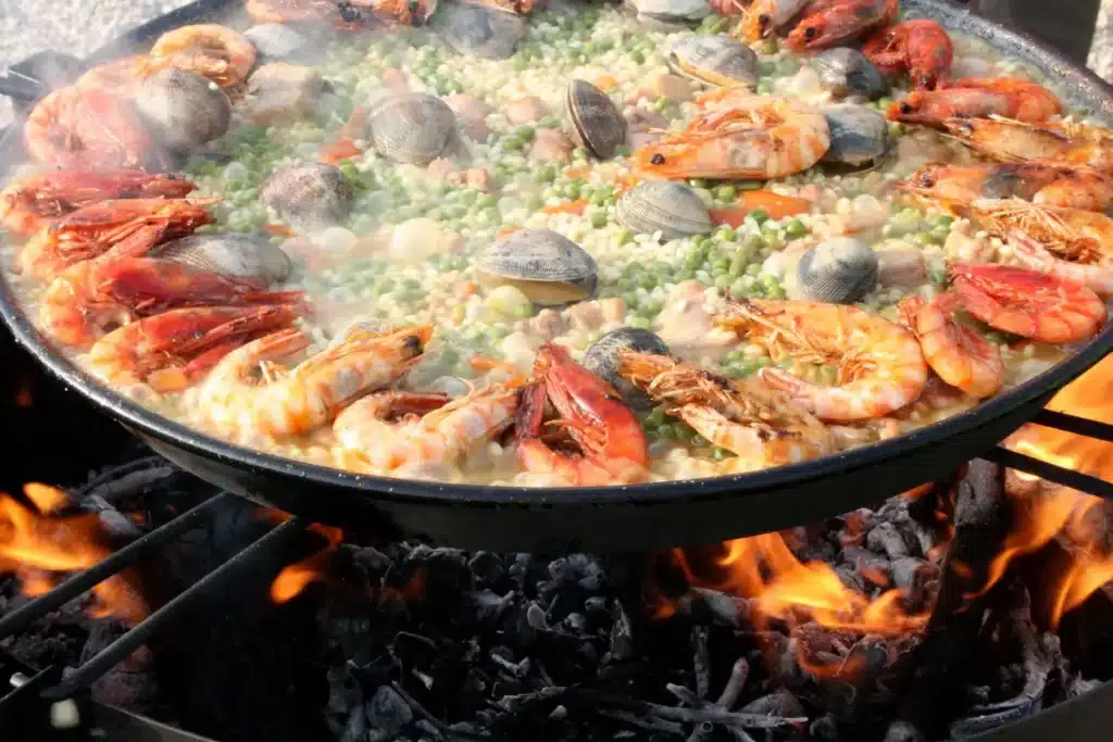 Eine Paellapfanne über einer offenen Flamme, gefüllt mit Garnelen, Muscheln, Erbsen und Reis, die auf einer Segelyacht gekocht wird, ist ein farbenfrohes und traditionelles Meeresfrüchtegericht.