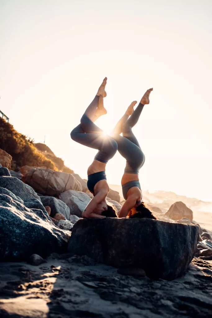 Zwei Menschen praktizieren bei Sonnenuntergang Yoga an einem felsigen Strand in der Nähe einer Segelyacht. Sie nehmen eine ausgeglichene Pose ein, wobei die Beine in einer Linie zum Himmel gestreckt sind und die Sonne durch ihre Silhouetten scheint.