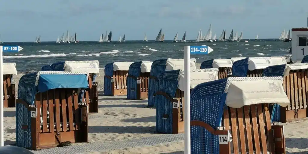 Reihen überdachter Strandstühle am Sandstrand mit Blick auf das Meer und zahlreiche Segelboote, die im Hintergrund unter einem bewölkten Himmel um die Wette schwimmen.