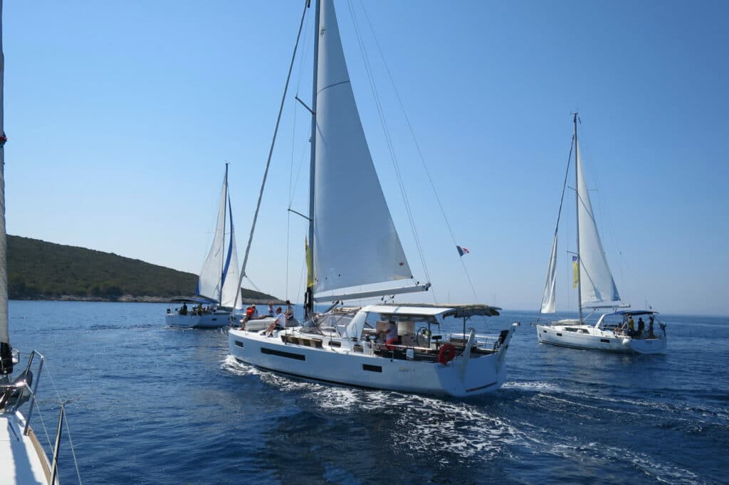 Mehrere Segelboote mit gesetzten Segeln gleiten an einem sonnigen Tag mit klarem Himmel und Hügeln im Hintergrund über ein ruhiges blaues Meer, ideal für einen Segelurlaub.