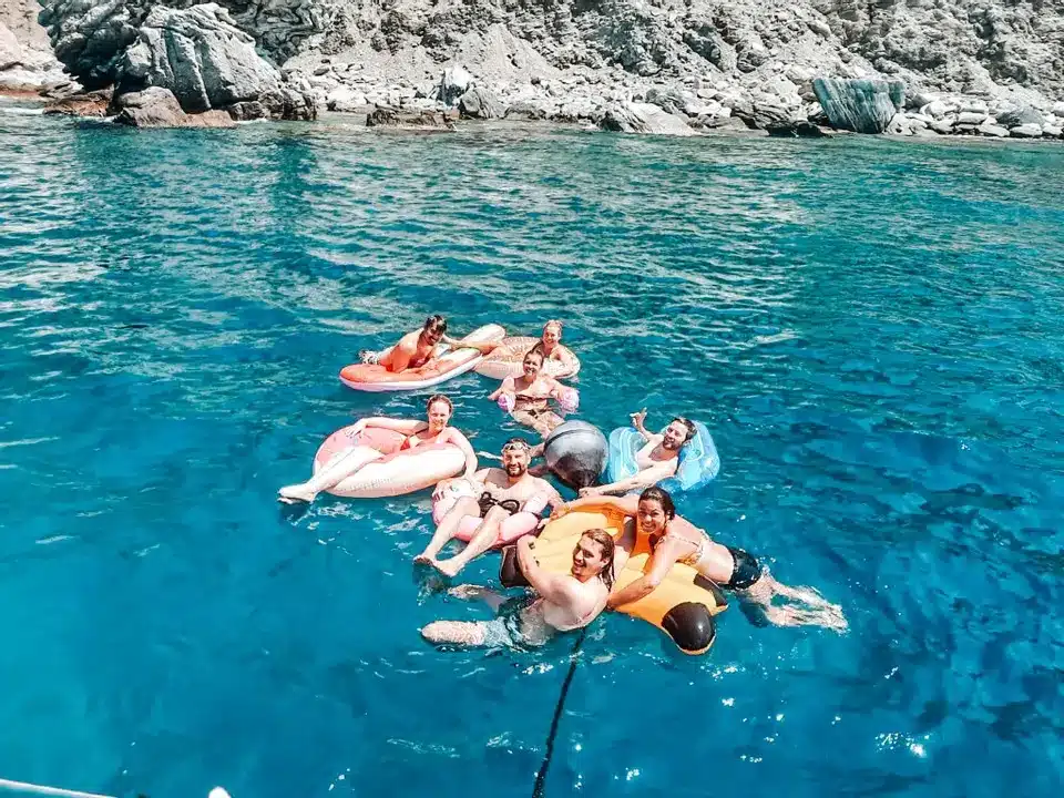 Eine Gruppe von Menschen treibt während ihres Segelurlaubs fröhlich auf bunten aufblasbaren Ringen im klaren blauen Wasser in der Nähe von felsigen Klippen.