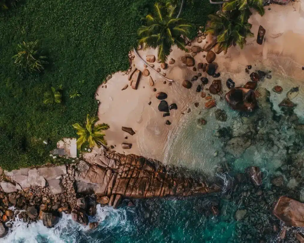 Luftaufnahme eines tropischen Strandes mit klarem, türkisfarbenem Wasser, felsiger Küstenlinie und üppigen grünen Palmen, der ein ruhiges und abgeschiedenes Segelurlaubsparadies zeigt.