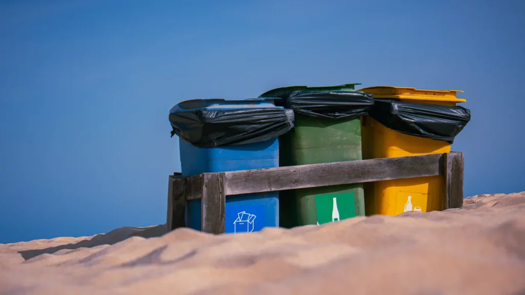 Drei bunte Recyclingbehälter (blau, grün und gelb) mit Deckeln, die mit schwarzen Säcken bedeckt sind, werden in einer Holzkiste an einem Sandstrand unter klarem Himmel in der Nähe einer Segelyacht gelagert.