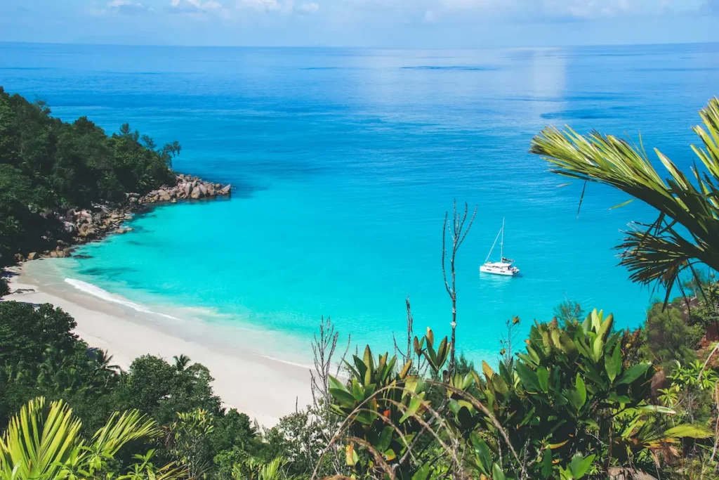 Ein ruhiger Strand mit türkisfarbenem Wasser, mit einem weißen Segelboot in Ufernähe, umgeben von üppigem Grün unter einem klaren blauen Himmel, perfekt für eine Segelreise.