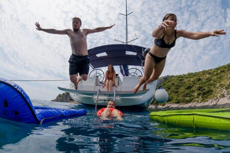 Zwei Menschen springen freudig von einer aufblasbaren Wasserrutsche neben einer Segelyacht ins Meer, während eine andere Person zusieht. Eine vierte Person schwimmt in der Nähe und genießt den sonnigen Tag.