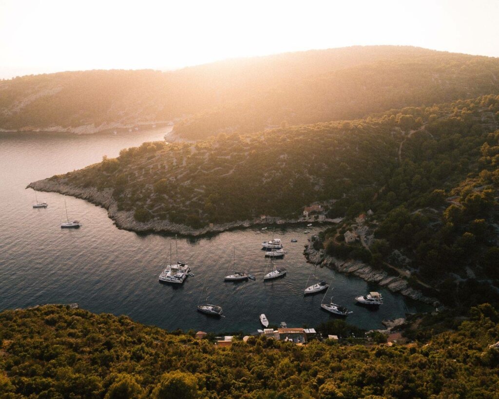 Luftaufnahme einer ruhigen Bucht mit mehreren Segelyachten, die in ruhigen Gewässern in der Nähe einer üppigen, bewaldeten Küste vor Anker liegen und im warmen Schein eines Sonnenuntergangs getaucht sind.