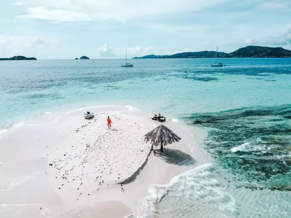 Luftaufnahme einer schmalen weißen Sandbank, die sich in türkisfarbenes Wasser erstreckt, mit einer einsamen Person in Rot, einem kleinen Strohschirm und zwei Jetskis. Üppige Hügel und eine Segelyacht