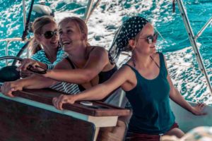 Drei Frauen lächeln und haben Spaß beim Steuern einer Segelyacht. Eine trägt einen Hut und eine Sonnenbrille. Sie sind vom blauen Meer umgeben.
