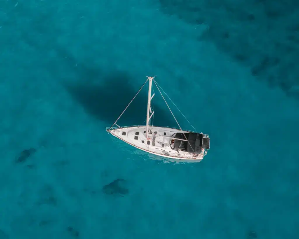 Luftaufnahme einer einsamen weißen Yacht, die auf einem leuchtend türkisfarbenen Meer segelt. Die Übersicht hebt die Isolation und die kontrastierenden Farben des Bootes und des Wassers hervor. Seitenwind
