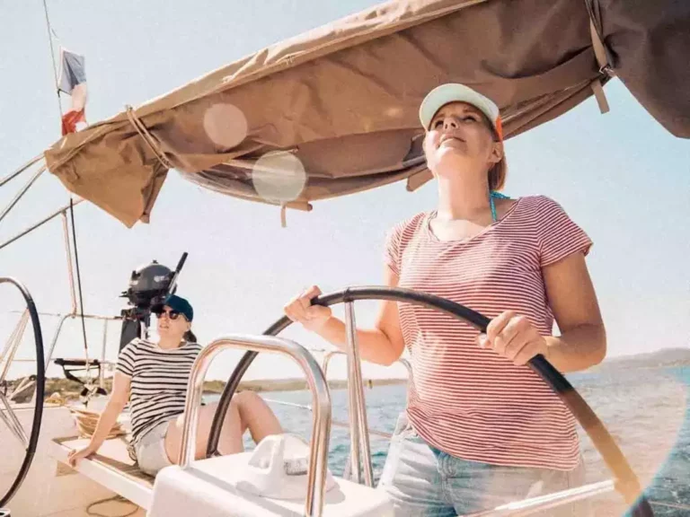 Eine Frau mit Mütze und gestreiftem Hemd steuert selbstbewusst ein Segelboot auf ihrer Segelreise, während im Hintergrund in einem schattigen Bereich eine weitere Person zu sehen ist.