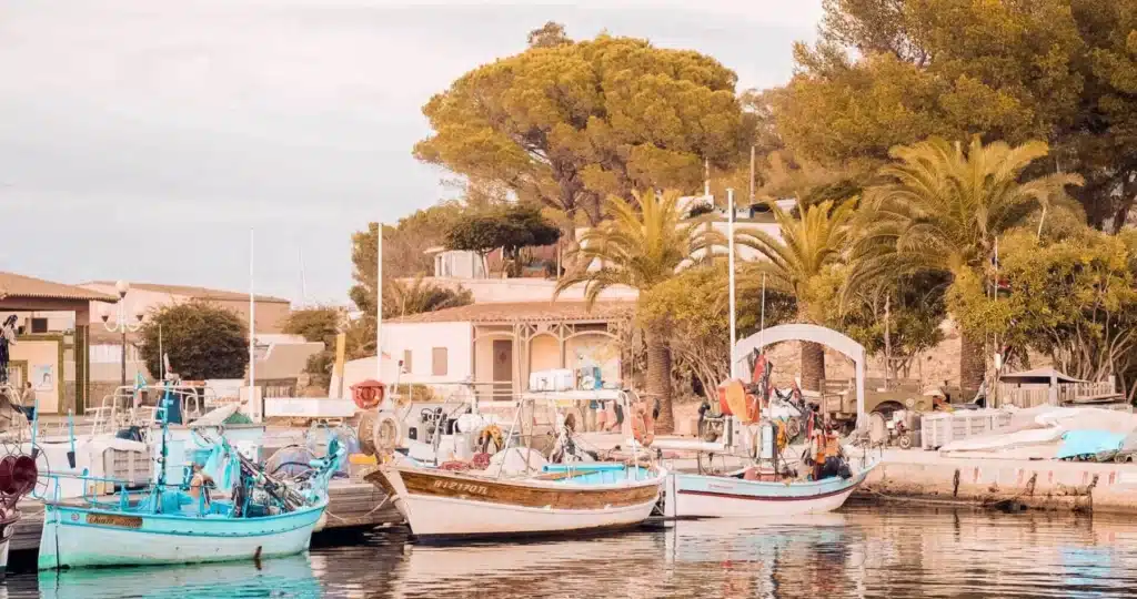 Eine ruhige mediterrane Hafenszene mit Segelyachten, die am Dock vertäut sind, flankiert von Palmen und kleinen Gebäuden unter einem sanften, warmen Himmel.