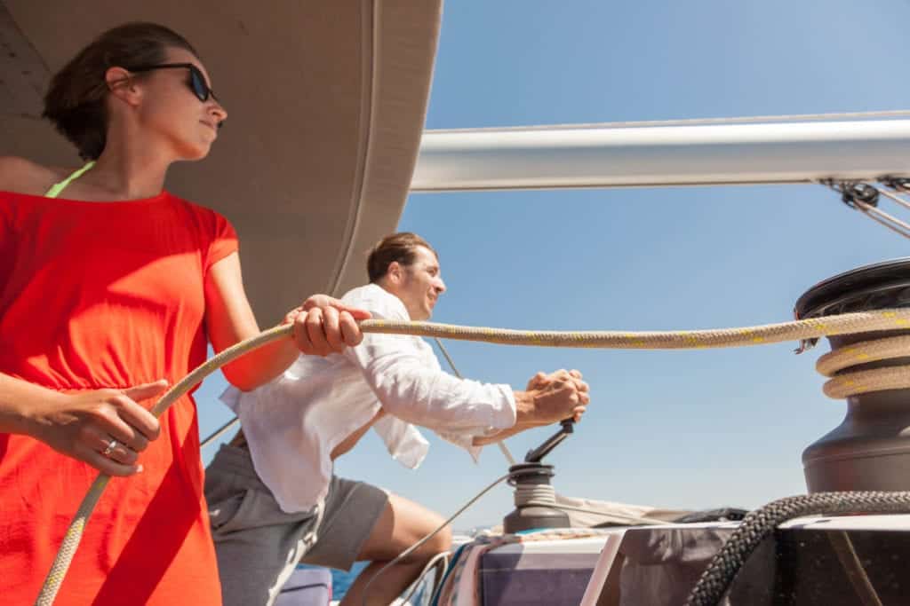 Zwei Personen auf einem Segelboot während eines Segeltörns: ein Mann im weißen Hemd, der ein Seil auf einer Winde aufwickelt, und eine Frau im roten Kleid, die zum Horizont blickt.