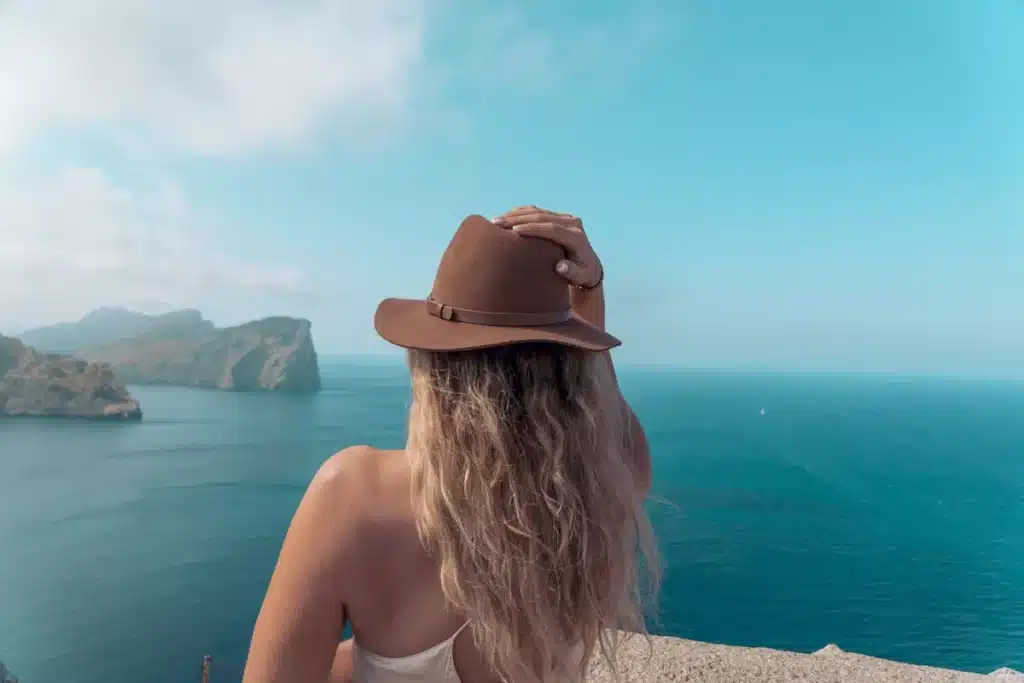 Eine Frau mit Hut blickt von einem hohen Aussichtspunkt an der Küste auf das ruhige blaue Meer und ist bereit, ihren Segeltörn zu starten. Darüber ist der Himmel dunstig blau und in der Ferne sind Klippen zu sehen.