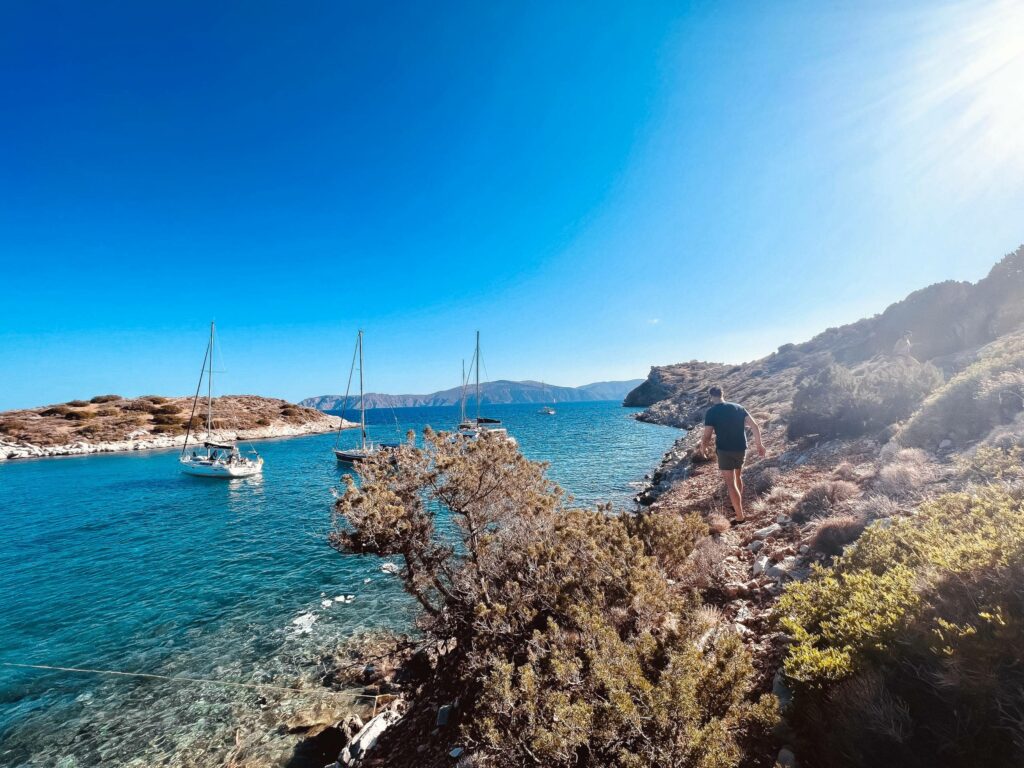 Eine Person steht während ihres Segelurlaubs an einer felsigen Küste mit Blick auf das klare blaue Meer, in dessen Nähe mehrere Segelboote vor Anker liegen, unter einem strahlend blauen Himmel, durch den die Sonnenstrahlen scheinen.