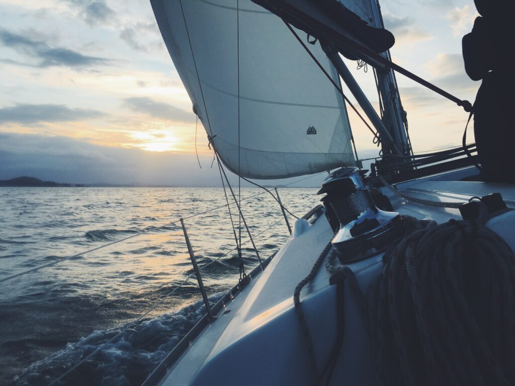 Blick von einer Segelyacht bei Sonnenuntergang. Das Deck, die Seile und die Segel des Bootes sind vor der Kulisse eines sanft gekräuselten Ozeans und eines bewölkten Himmels zu sehen, der von der untergehenden Sonne beleuchtet wird.