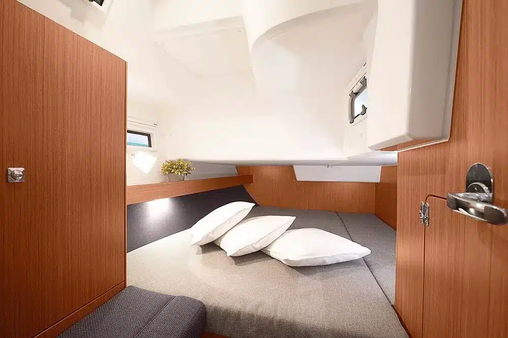 Kompakte Segelyachtkabine mit Holzwänden, einem Bett mit Kissen, einem kleinen Fenster und einem grauen Teppichboden. Auf der rechten Seite ist eine Tür angelehnt, was auf mehr Innenraum hindeutet