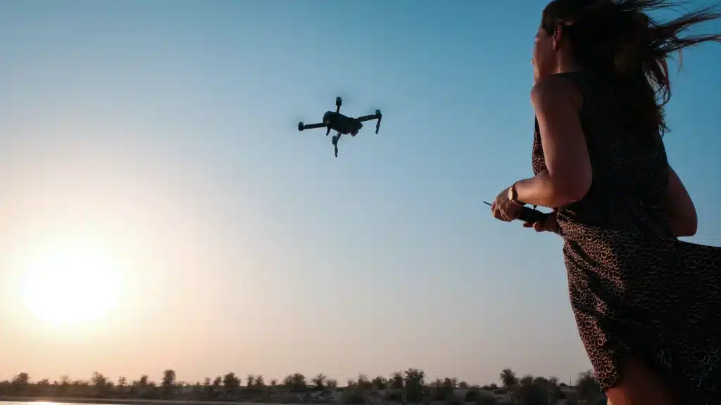 Eine Frau in einem Kleid mit Leopardenmuster steuert bei Sonnenuntergang eine Drohne. Die Drohne fliegt durch den Himmel und im Hintergrund ist die Sonne am Horizont zu sehen.