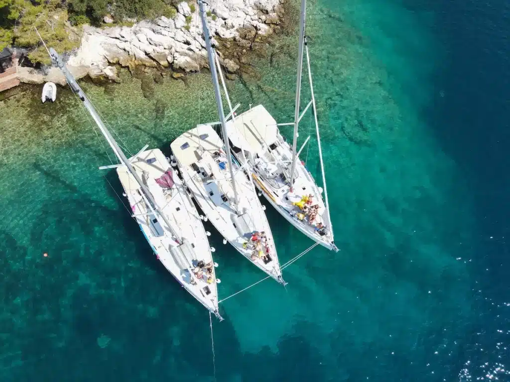 Luftaufnahme von drei Segelbooten, darunter ein Katamaran, die dicht beieinander in der Nähe einer felsigen Küste vertäut sind. Klares blaugrünes Wasser umgibt sie und betont die natürliche Schönheit des Ortes.