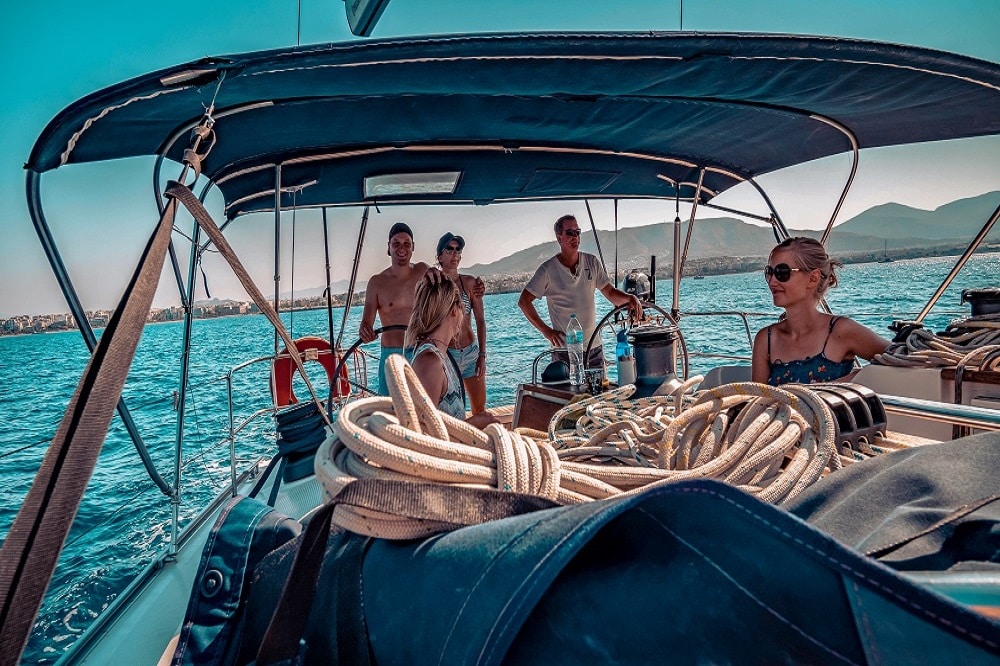 Vier Menschen genießen einen sonnigen Tag auf einem Segeltörn, umgeben von Seilen und Segelausrüstung, mit einem klaren blauen Himmel und einer bergigen Küste im Hintergrund.