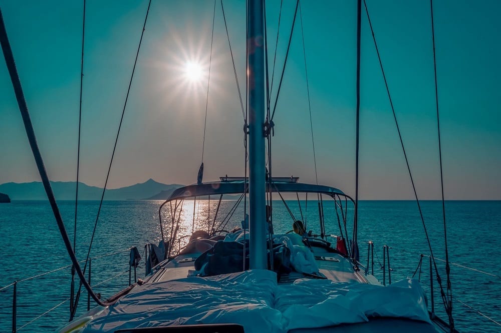 Blick vom Deck einer Segelyacht in Richtung der über dem Meer untergehenden Sonne. Masten und Takelage sind als Silhouetten vor einem strahlend blauen Himmel zu sehen.