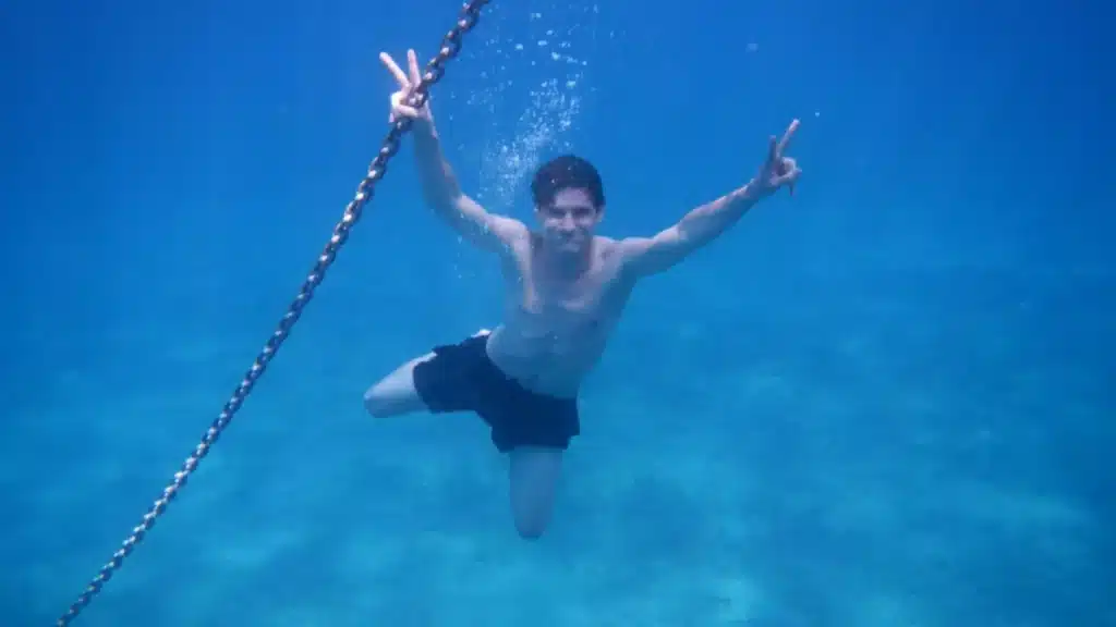 Ein Mann macht unter Wasser mit beiden Händen ein Peace-Zeichen, während er sich an einer vertikalen Kette festhält und im klaren blauen Wasser in der Nähe einer Segelyacht liegt.