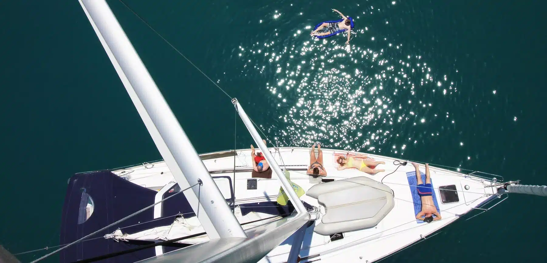 Luftaufnahme einer Yacht auf klarem, blauem Wasser mit Menschen, die auf dem Deck ein Sonnenbad nehmen, und einer Person, die während ihres Segelurlaubs vom Mast ins Wasser springt.