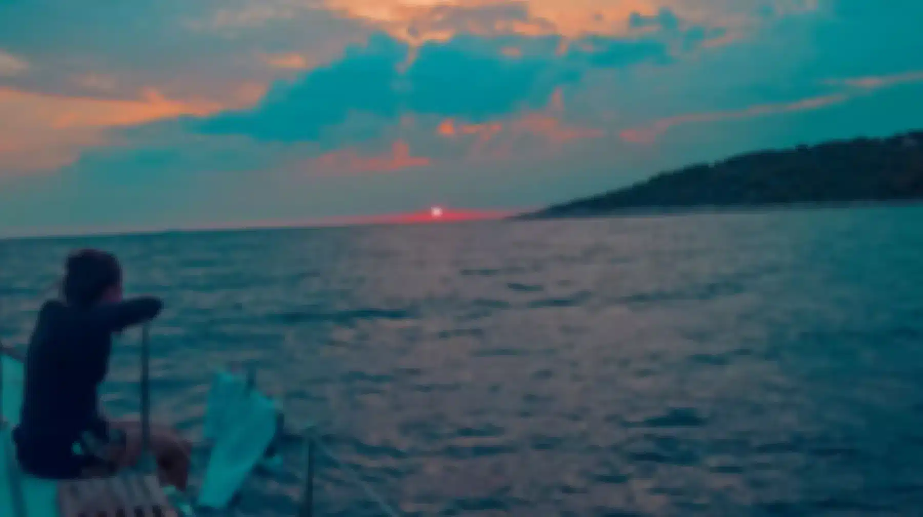 Eine Person, die auf der Rückseite eines Bootes sitzt und während einer Segelreise einen Sonnenuntergang über einem ruhigen Meer beobachtet, mit einer teilweise sichtbaren Sonne, die in der Nähe eines hügeligen Horizonts unter einer lebhaften Wolke untergeht