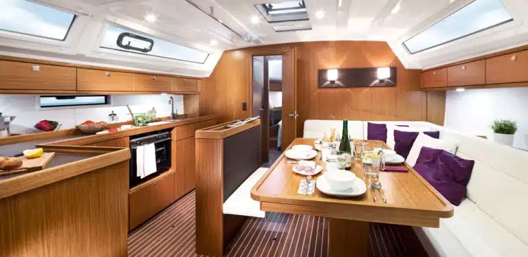 Das Interieur einer luxuriösen Yacht mit einer gut ausgestatteten Küche auf der linken Seite und einem Essbereich für vier Personen auf der rechten Seite, umgeben von eleganten Holzoberflächen und natürlichem Licht, bereit für einen Segeltörn.